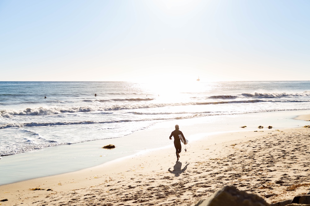 Surfer in wetsuit running toward the ocean waves on a sandy beach under clear sky at sunrise or sunset.