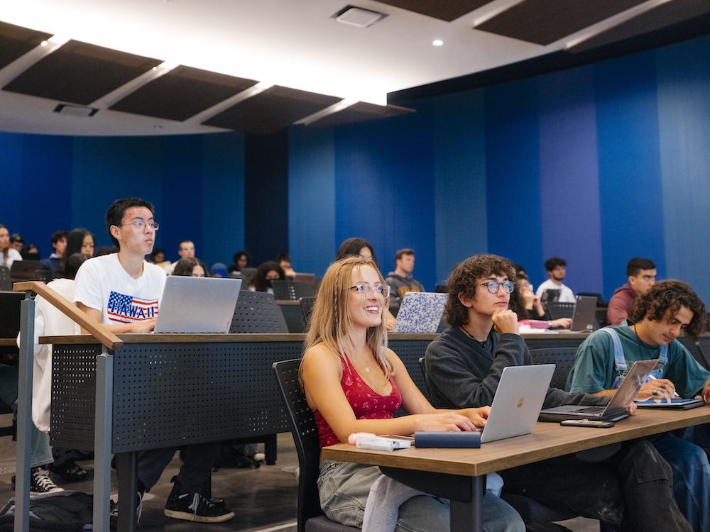 Students sit at desks with laptops in a lecture hall with blue walls, engaged in a class or workshop setting with dozens of participants visible.