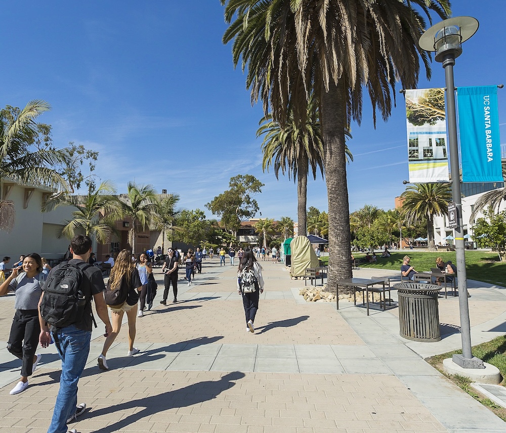UCSB campus plaza on a sunny day with students walking beneath tall palm trees, campus buildings visible in background, and a turquoise "SANTA BARBARA" banner hanging from a street lamp.