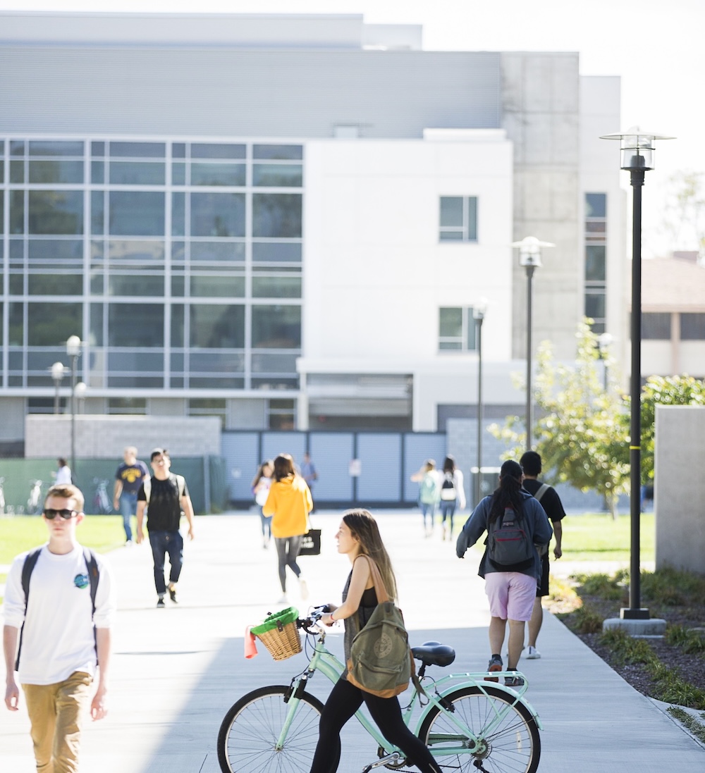 Students walk and bike across a sunny campus plaza near modern buildings. A student in the foreground rides a light green bike with a basket, while others walk in the background on a paved courtyard.