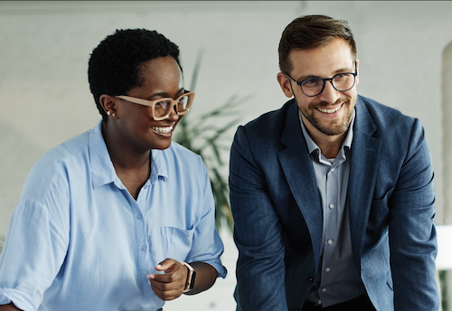 Two professionals both wearing glasses in dress attire smiling in an office setting. 
