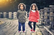 Two young children standing on a wooden boardwalk surrounded by white fencing. Child on left wears olive jacket and blue jeans. Child on right wears red jacket with white collar and black leggings with red shoes.
