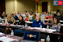 Group of diverse professionals seated at conference tables in a classroom setting, smiling and engaged during a training or workshop session, with water bottles and materials on tables.
