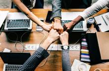 Four professionals place their fists together in a team unity gesture over a wooden desk with laptops, phones, and office supplies.