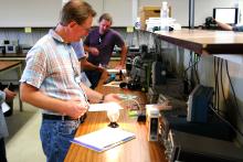 Instructor wearing glasses and striped shirt demonstrates infrared equipment to participants in a laboratory classroom setting with 3D printer and monitoring displays visible.
