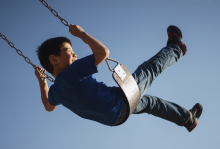 Child on swing against clear blue sky, wearing dark blue shirt and jeans, legs extended outward in mid-swing motion.