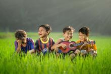 Four children sit in a grassy field, smiling while one plays a red acoustic guitar. Overcast sky visible in background.