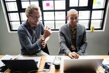 Two professionals collaborating at a wooden desk with laptops and mobile devices in a modern office with large windows and sticky notes on glass.