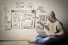 Person sitting against wall with laptop, sketching website wireframe on wall with pen, showing user interface layout design with boxes and navigation elements.